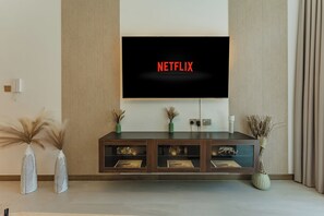 Ett modernt vardagsrum med en platt-TV som är monterad på väggen och visar "NETFLIX", ett träinrett TV-bord nedanför med dekorativa föremål, och två höga vaser med torkade växter.