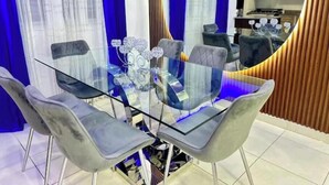 Un comedor moderno con una mesa de vidrio y sillas grises tapizadas, un jarrón decorativo y una cocina al fondo.