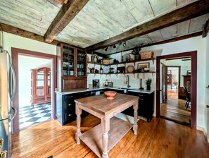 A rustic kitchen with wooden beams, a central table, and shelves with kitchenware. - Elegant Luxury Cottage with Fire Pit Near Carrying Place Beach in Surry, Maine (Surry)
