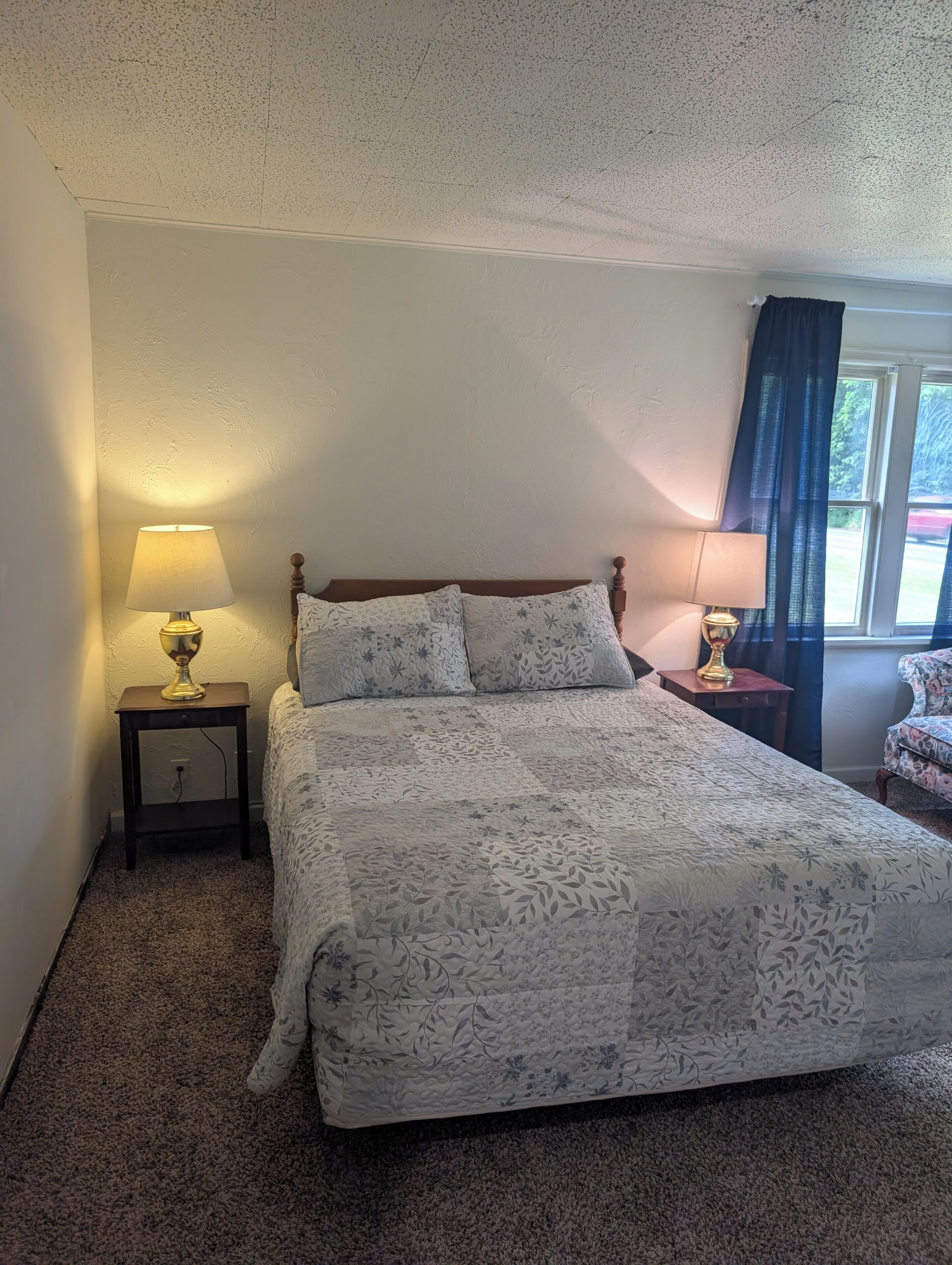 A bedroom with a bed, two bedside lamps, a window with curtains, and a carpeted floor.