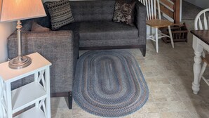 Un escalier avec des rampes en bois, un canapé gris avec des coussins à motifs, une petite table blanche avec une lampe et un tapis tissé sur un sol carrelé.