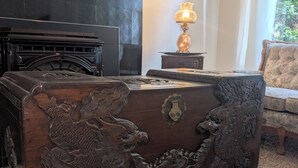 Un coffre en bois traditionnel orné de sculptures complexes, une cheminée, une lampe et un canapé à motifs dans une pièce dont le sol est recouvert d’une moquette.