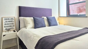 Uma cama bem arrumada com cabeceira, uma mesa de cabeceira com uma placa e uma janela com vista para uma parede de tijolos.