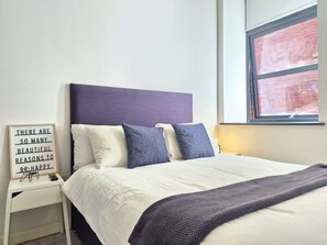 A neatly made bed with a headboard, a bedside table with a sign, and a window with a view of a brick wall. - Chic Industrial Loft-Style Apartment,Central Leeds (Leeds)