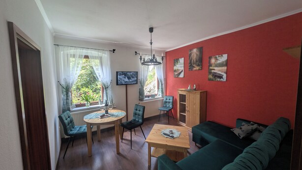 A living room with a red wall, a wooden cabinet, a green sofa, a dining table with chairs, and a chandelier. - Ulbersdorf Mill "Försterstübchen" (Hohnstein)