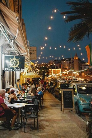 A bustling outdoor dining area with tables and chairs, string lights overhead, and a sign for a bar and snack bar. - A 3BR equipped home w/WIFI Next to St Thomas Bay (Marsaskala)