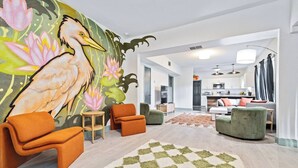 Uma sala de estar moderna com um mural, sofás laranja e verde, um tapete estampado e uma cozinha ao fundo.