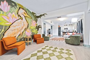 Ein modernes Wohnzimmer mit Wandgemälde, orangefarbenen und grünen Sofas, einem gemusterten Teppich und einer Küche im Hintergrund.