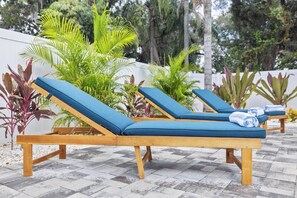 Zwei Holz-Loungesessel mit blauen Kissen stehen auf einer gefliesten Terrasse, im Hintergrund sind Kübelpflanzen zu sehen.