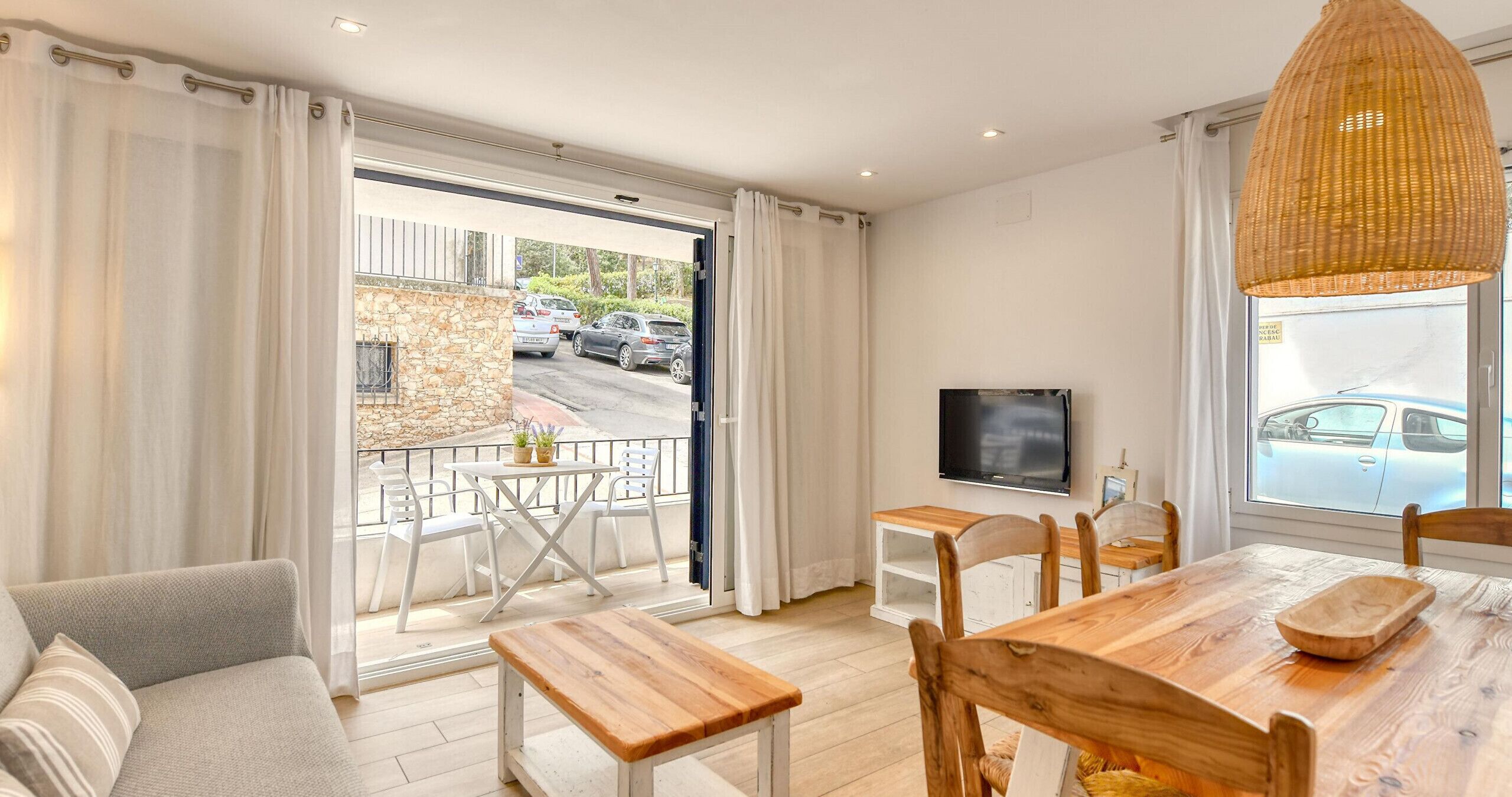A modern living room with a wooden dining table, white chairs, a sofa, a TV, and a balcony with a view of parked cars.