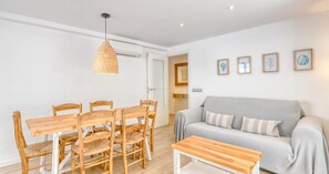 Ein Wohnzimmer mit grauem Sofa, Esstisch aus Holz, Korbstühlen und einer hängenden Leuchte, sowie gerahmten Kunstwerken an der Wand.