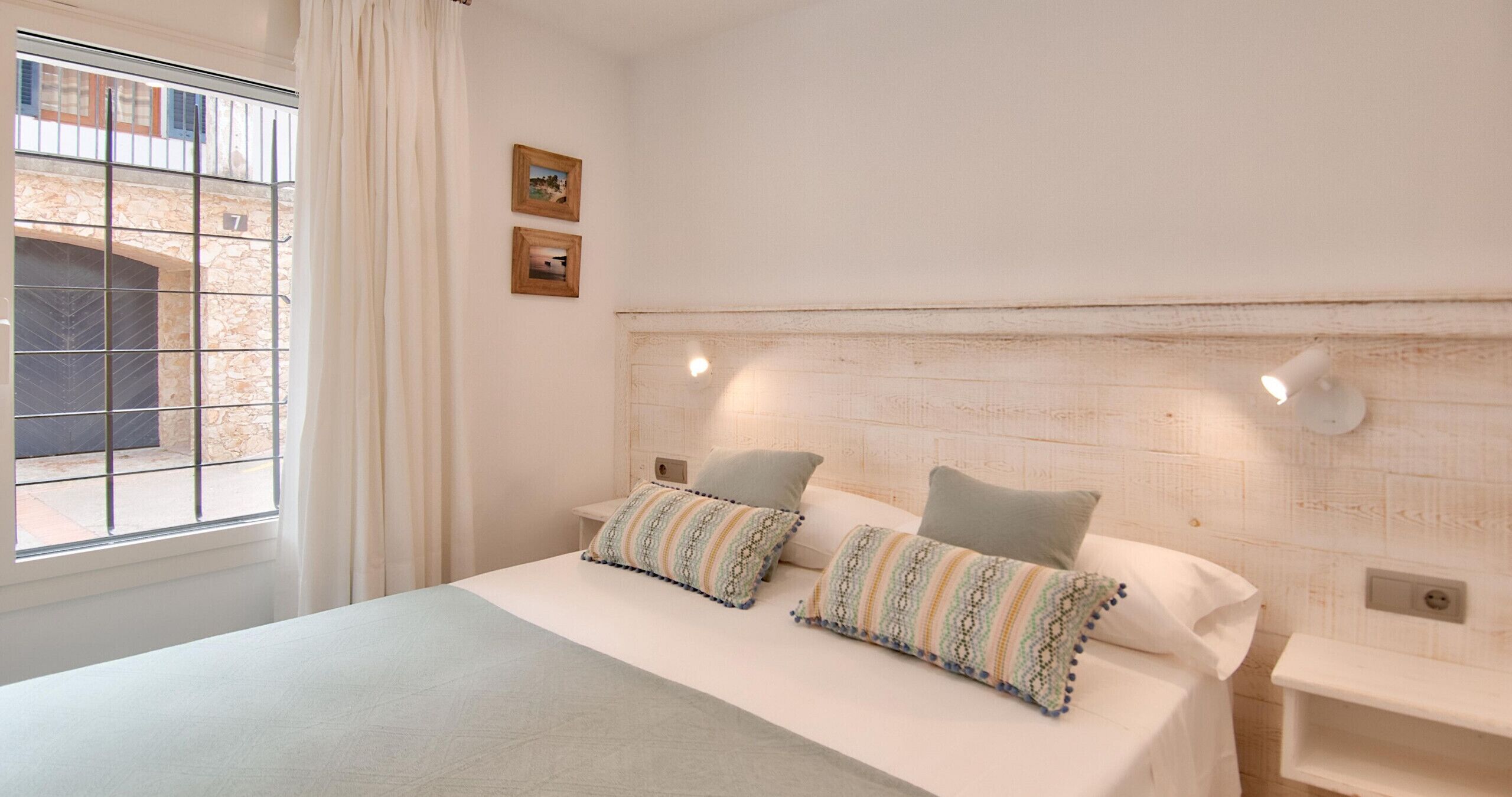 Ein Schlafzimmer mit einem Bett, zwei Kissen, einem Nachttisch, einem Fenster mit Vorhängen und einer Wandleuchte.