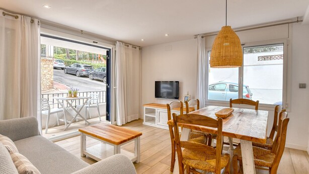 A modern living room with a sofa, dining table, chairs, a TV, and a balcony with a view of the street. - Renovated ground-floor apartment in the center of Calella de Palafrugell (Calella de Palafrugell)