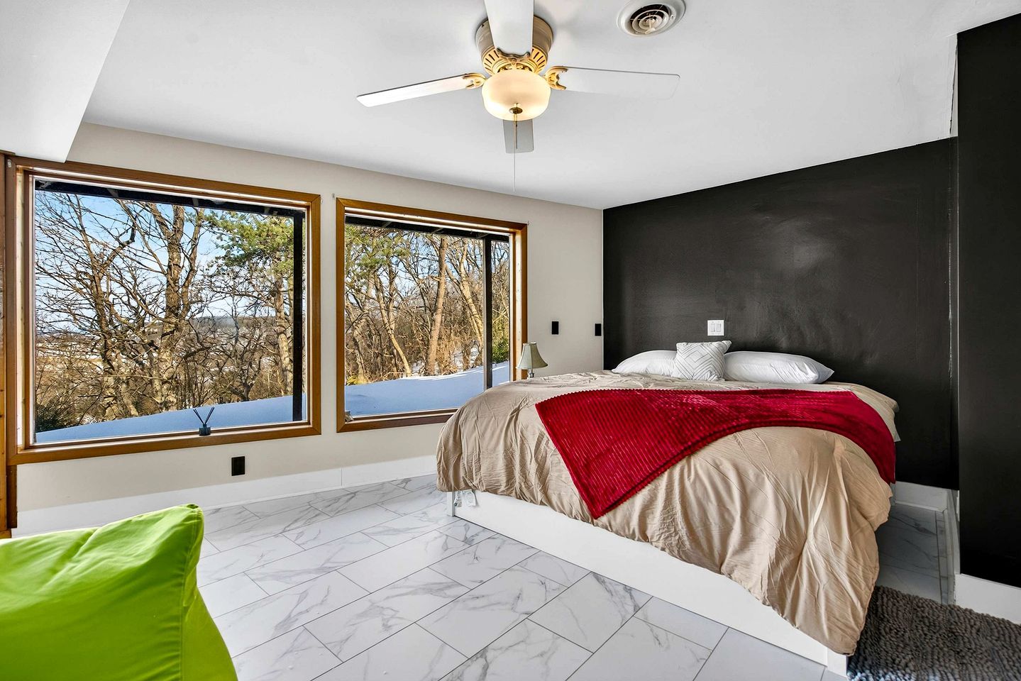 A bedroom with a bed, large windows, a ceiling fan, and a view of trees.