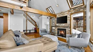 A spacious living room with a fireplace, a large TV, and a high ceiling with exposed beams.