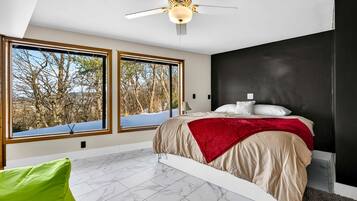 Une chambre à coucher avec un lit, de grandes fenêtres, un ventilateur de plafond et une vue sur les arbres.