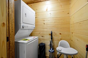 A compact bathroom with a washing machine, a vacuum cleaner, and a white chair. - Luxury Gatlinburg Cabin with Game Room & Hot Tub near Trolley in Gatlinburg, Tennessee (Gatlinburg)