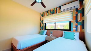 Ein Hotelzimmer mit zwei Betten, einem Deckenventilator, einer Klimaanlage, einem Fenster mit Jalousien und einem farbenfrohen Wandmuster.