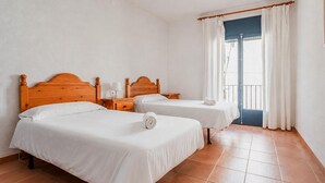 Habitació amb dos llits, un capçal de fusta, una petita taula de nit, una finestra amb cortines i una porta.