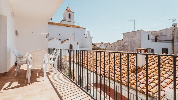 Un balcó amb cadires de plàstic blanc, un paviment de rajola i vista d'un edifici blanc amb teulada vermella i un campanar.
