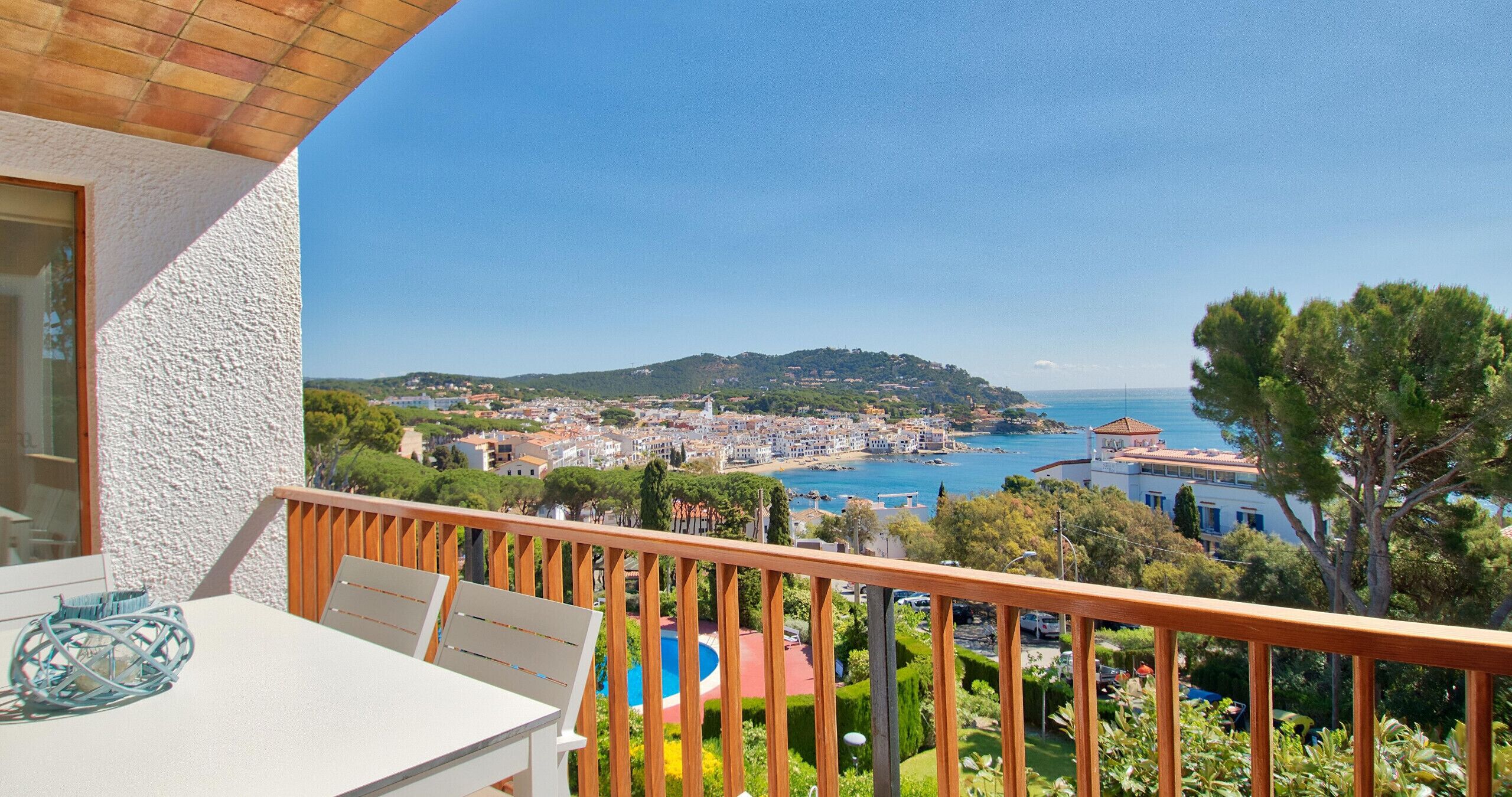 Ein Balkon mit Tisch und Stühlen, der auf eine Küstenstadt mit freiem Blick auf das Meer und eine hügelige Landschaft im Hintergrund blickt.