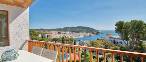A balcony with a table and chairs, overlooking a coastal town with a clear view of the sea and a hilly landscape in the distance.