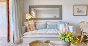 Ein gemütliches Wohnzimmer mit einer Couch, Kissen, einer Lampe, einem Korbstuhl und einer Vase mit Blumen.