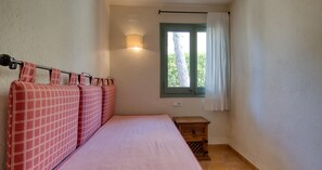 A small, single-bed room with a window, a wall-mounted lamp, and a small wooden nightstand.