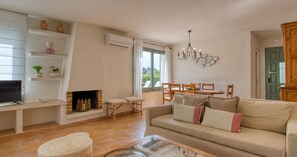 A living room with a sofa, TV, fireplace, dining table, chairs, and a chandelier. - 4-Bedroom Villa with Pool in Calella · Ideal for Families (Calella de Palafrugell)