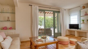 Una sala de estar con una mesa de centro de vidrio, muebles de mimbre, un televisor y una puerta corrediza de vidrio que da a un espacio exterior.