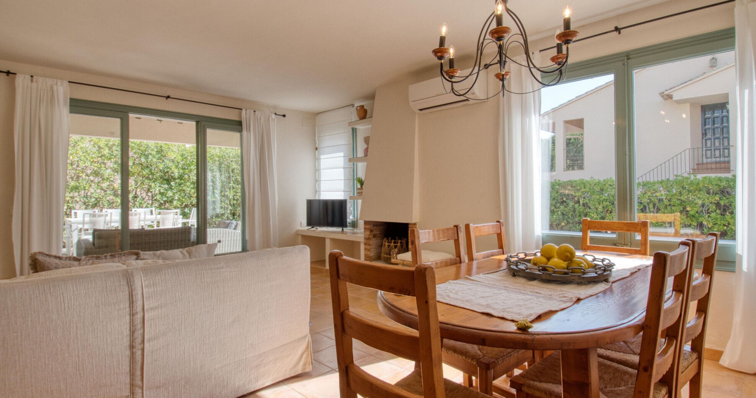 Een eetruimte met een houten tafel, stoelen, een kroonluchter en uitzicht op groen.