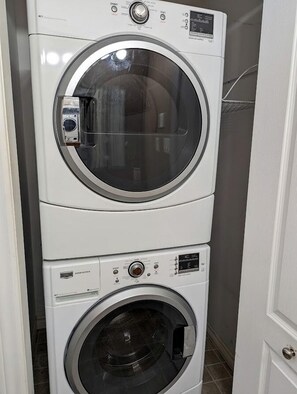 A stacked washer and dryer unit in a room with a door and tiled floor. - Spacious Ranch-Style Retreat with Private Deck, Nestled in Nature Near Acadia, Hancock Maine (Hancock)