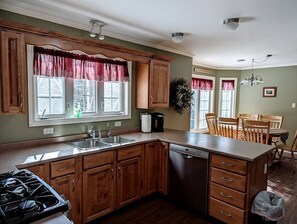A kitchen with wooden cabinets, a dishwasher, and a dining area with a table and chairs. - Spacious Ranch-Style Retreat with Private Deck, Nestled in Nature Near Acadia, Hancock Maine (Hancock)