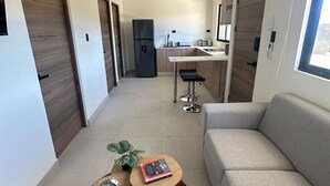 A modern living room with a sofa, a small round table with a book, a potted plant, and a kitchen area with a refrigerator and sink in the background. - Tempátika Villa 3, New (Tempate)