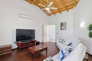 Sala de estar con televisor de pantalla plana, mueble de madera para entretenimiento, sofá blanco con cojines azules y ventilador de techo.