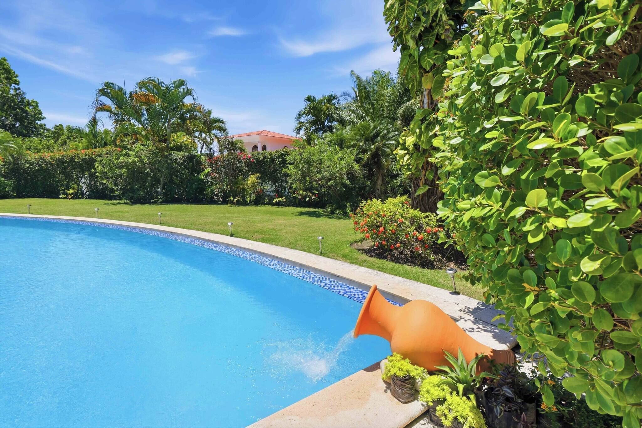 A swimming pool with a tiled edge, surrounded by lush greenery and a building in the background.