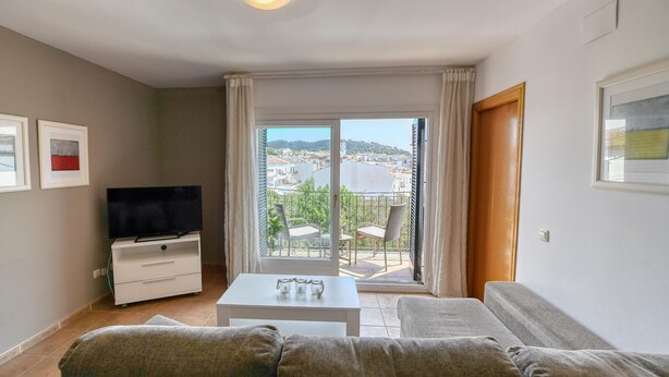 Ein Wohnzimmer mit einer Couch, einem Fernseher, einem weißen Couchtisch und einem Balkon mit Blick auf eine Stadt und Wasser.