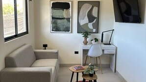Ein modernes Wohnzimmer mit einer Couch, einem Schreibtisch, einem Sessel, einem kleinen Tisch mit Pflanze und Kunstwerken an den Wänden.