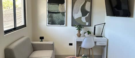 A modern living room with a sofa, a desk, a chair, a small table with a plant, and artwork on the walls.