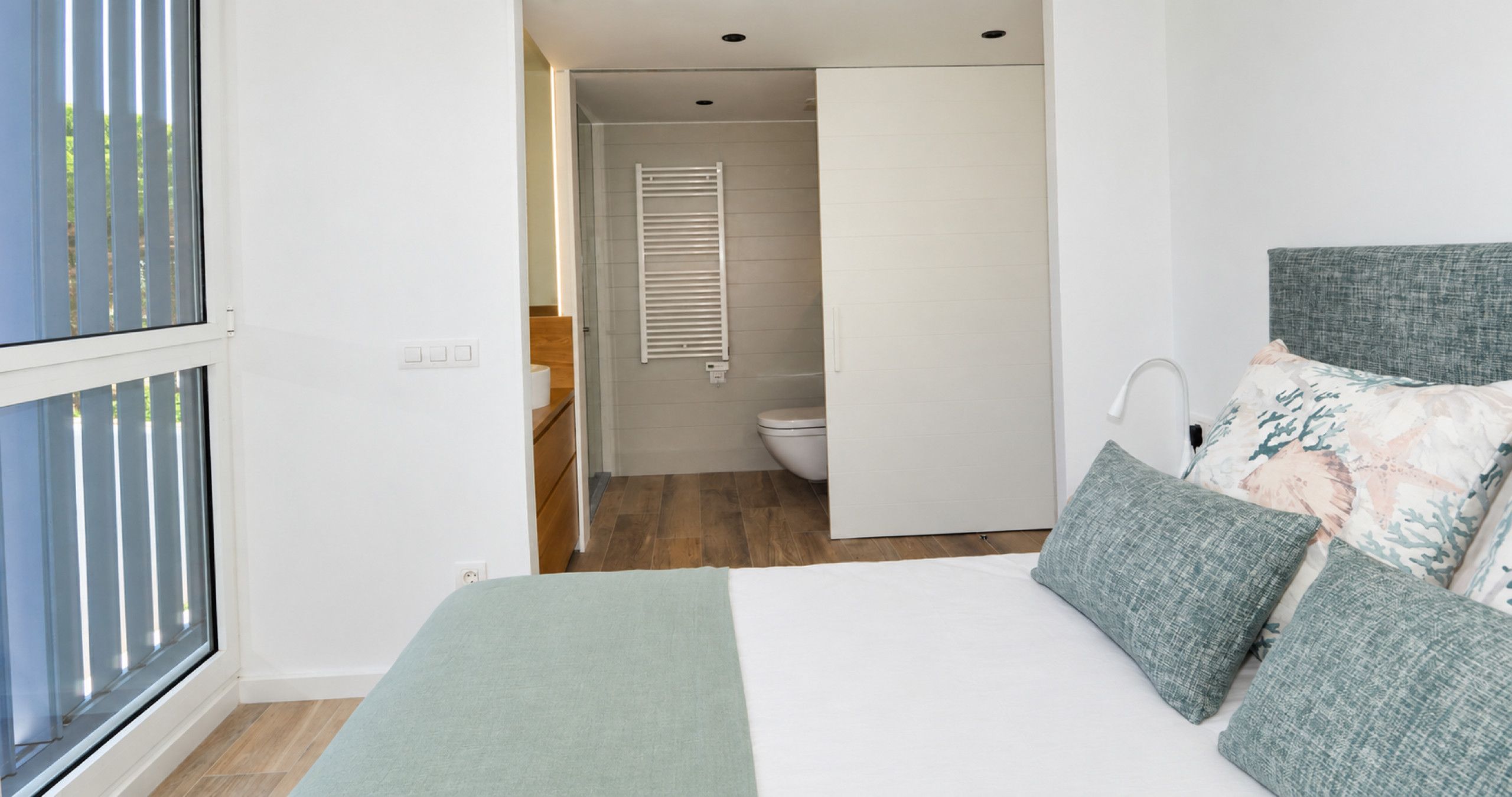 Une chambre à coucher avec un lit, des oreillers, une table de chevet, une fenêtre et une vue sur la salle de bain.