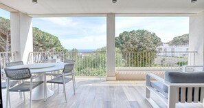 Uma varanda com mesa e cadeiras, um sofá e vista para árvores e o mar.