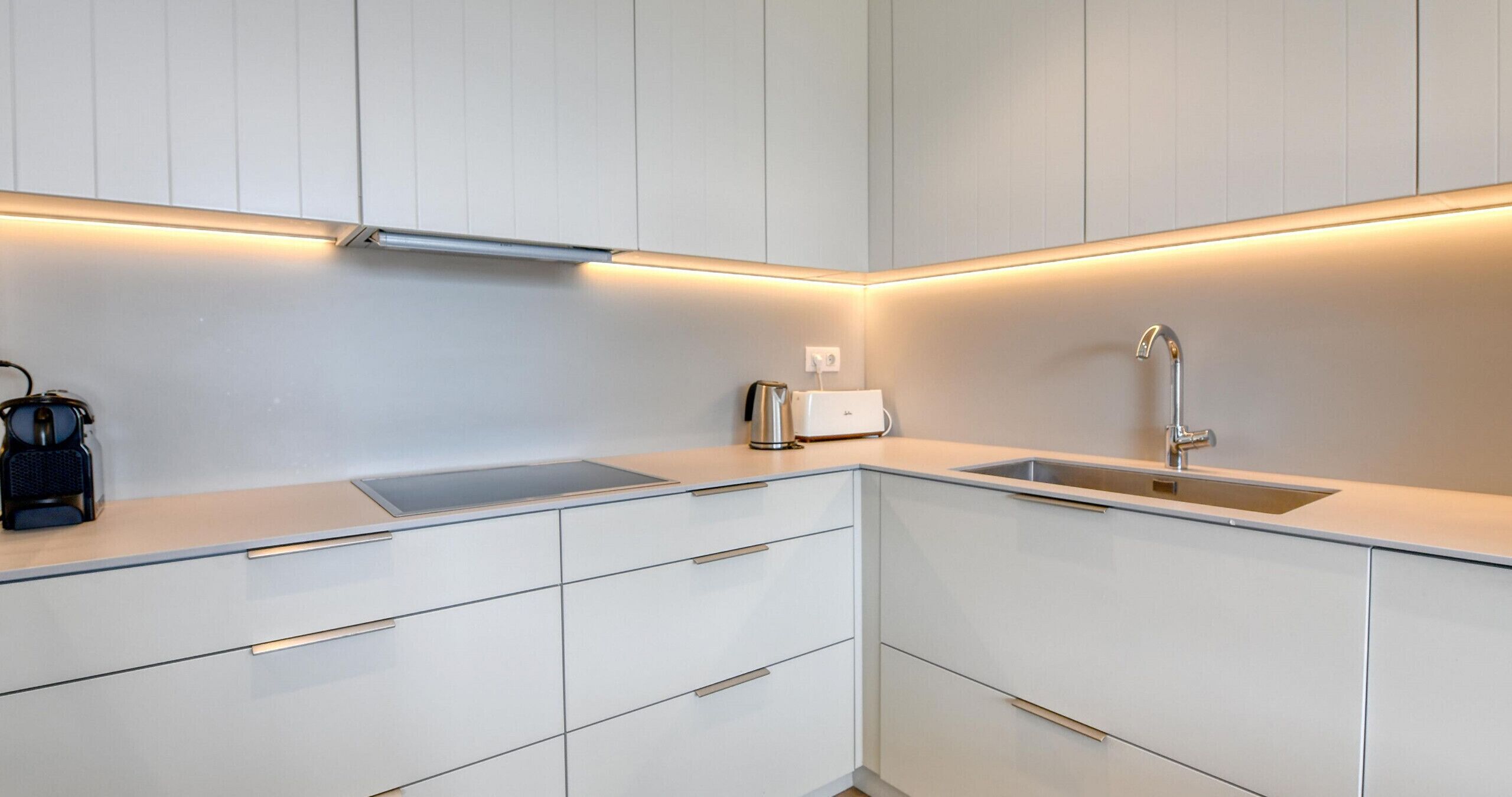 Une cuisine moderne avec des meubles blancs, un évier en acier inoxydable et une bouilloire posée sur le plan de travail.