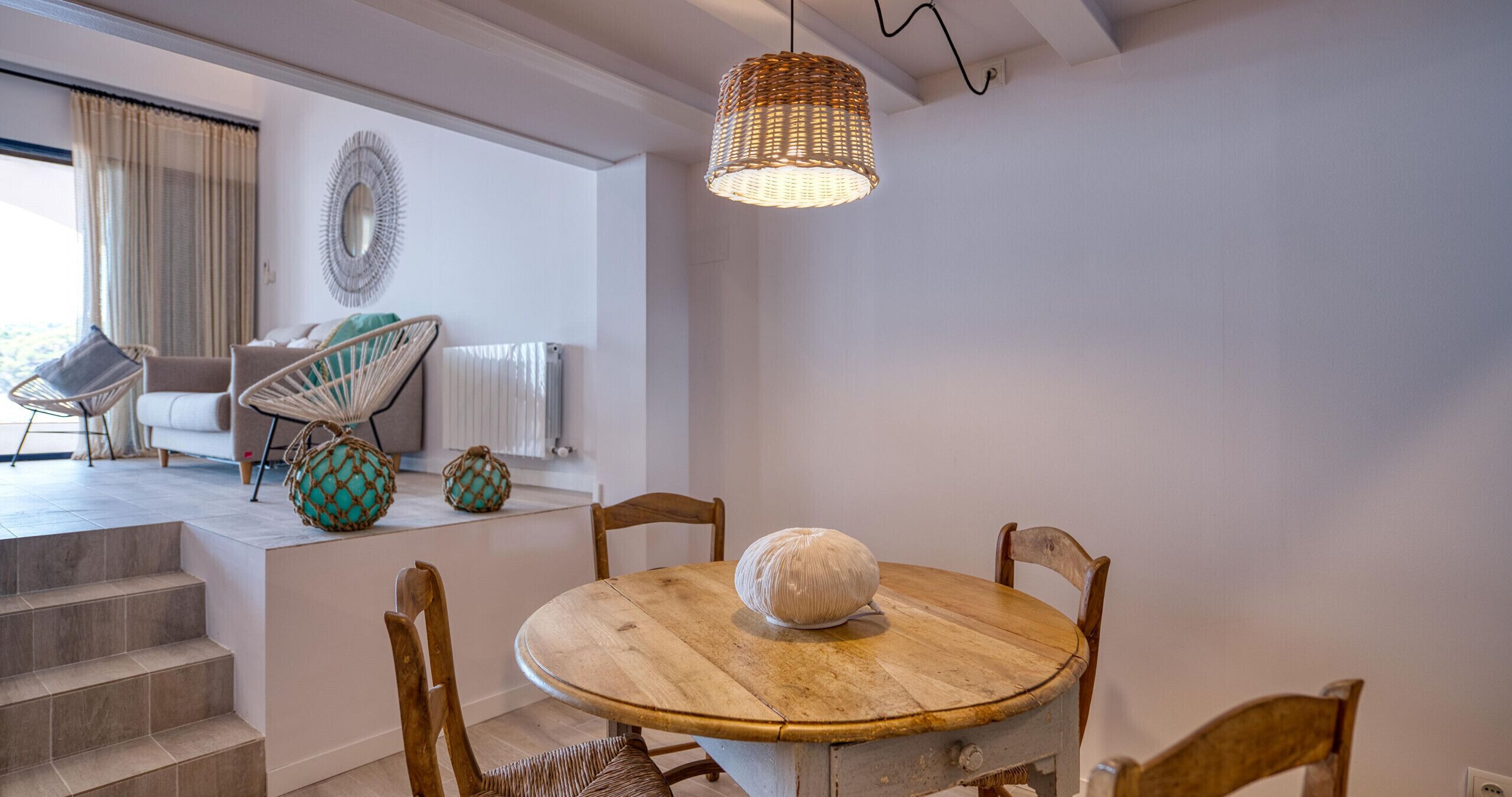 Un coin repas comprenant une table ronde en bois, quatre chaises, un luminaire suspendu en fibres tressées et un miroir décoratif accroché au mur.