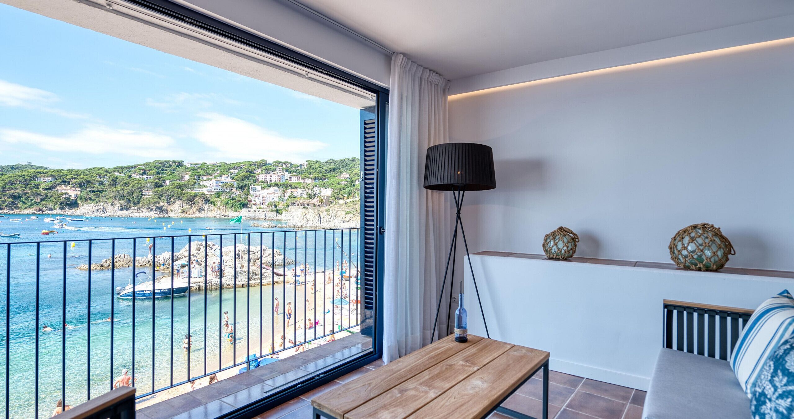 Un balcon avec vue sur la mer et sur le paysage urbain, une table en bois, une lampe et des objets décoratifs sur une étagère.