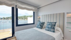 Un dormitorio con un ventanal grande, una cama con almohadones estampados, una mesita de noche con lámpara y vista al mar.
