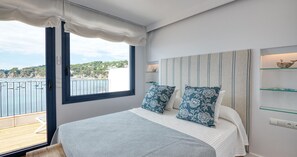 Ein Schlafzimmer mit großem Fenster, einem Bett mit gemusterten Kissen, einem Nachttisch mit Lampe und Blick auf das Meer.