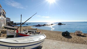 Escena costera con barcos amarrados en una playa de arena, cielo despejado y un promontorio rocoso en el agua.