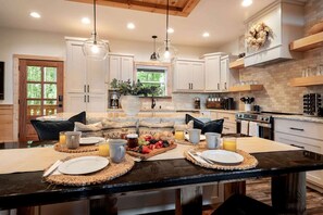 A modern kitchen with white cabinets, a black countertop, and a breakfast setup with fruits and beverages. - Luxury Mountain Cabin with Hot Tub, Sauna & Game Room Near Ellijay in Georgia (Ellijay)