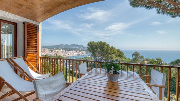 Ein Balkon mit einem Holztisch und zwei weißen Sitzgelegenheiten mit Kissen, mit Blick auf eine Küstenstadt und freiem Ausblick auf das Meer und die entfernten Hügel.