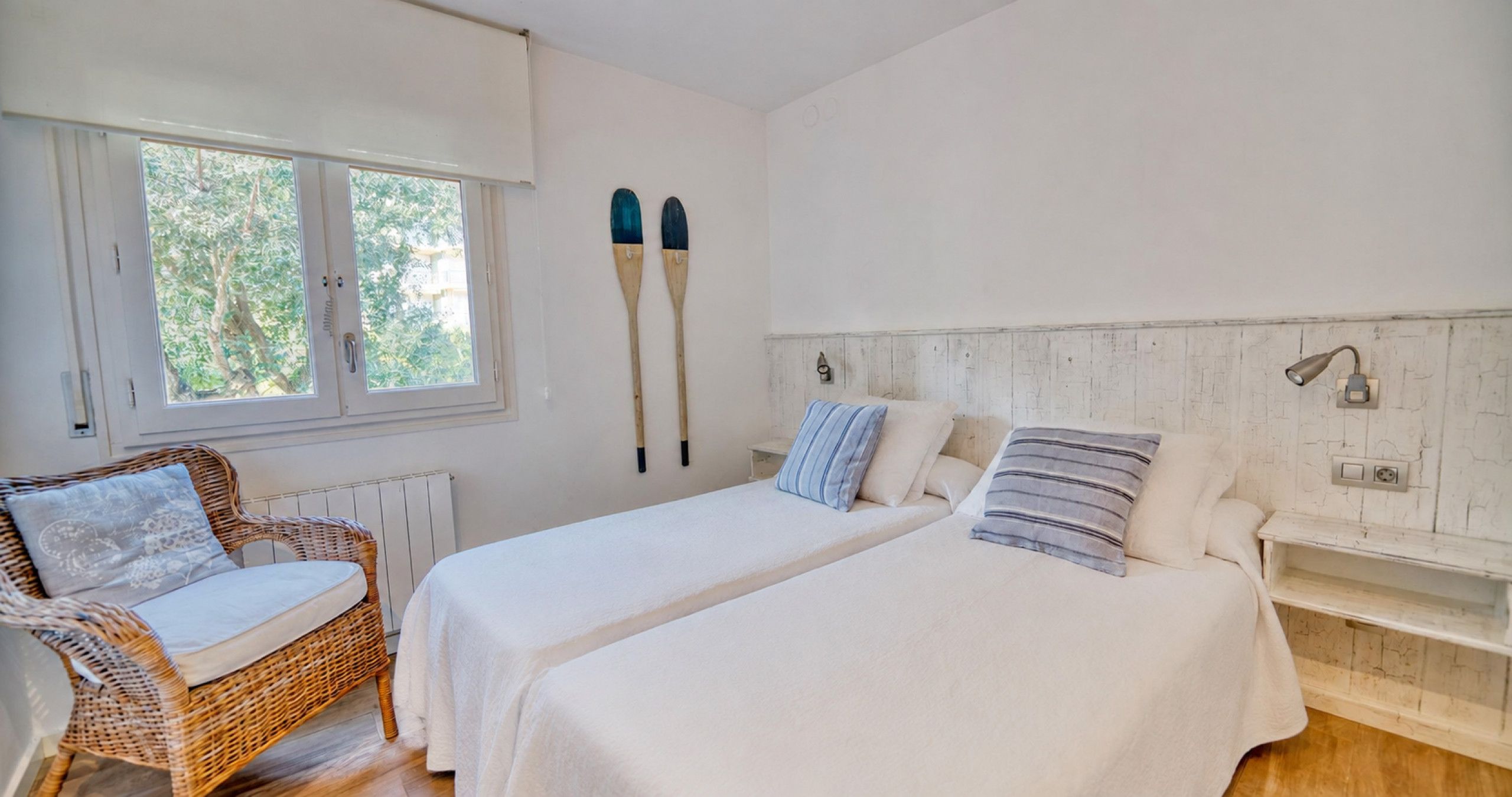 A bedroom with two beds, a wicker chair, a window with a view of greenery, and a wall-mounted light fixture.